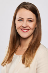 Nahaufnahme einer lächelnden Frau mit langen dunklen Haaren vor hellem Hintergrund.