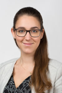Nahaufnahme einer lächelnden Frau mit Brille vor einem hellen Hintergrund.