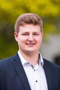 Portrait eines jungen Mannes im blauen Sakko und weißem Hemd vor grünem Hintergrund.