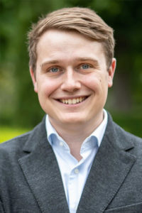 Portrait eines lächelnden jungen Mannes mit kurzen blonden Haaren in dunklem Sakko vor grünem Hintergrund.