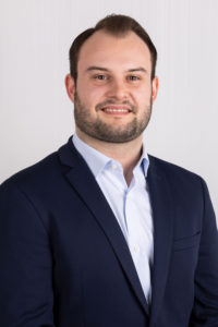 Portrait eines bärtigen Mannes im dunklen Sakko und weißem Hemd vor hellem Hintergrund.