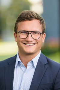 Portrait eines jungen Mannes mit Brille und Seitenscheitel im blauen Sakko vor unscharfem Hintergrund.