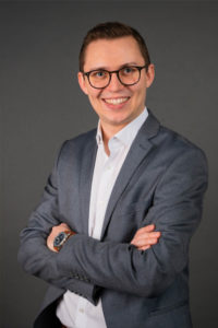Portrait eines jungen Mannes mit Brille im dunklen Sakko, Arme verschränkt, vor hellem Hintergrund.