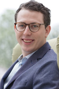 Portrait eines jungen Mannes mit Brille und kurzem Haar, lächelnd in Außenumgebung.