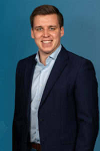 Portrait eines Mannes im dunklen Anzug und hellblauem Hemd vor dunklem Hintergrund.